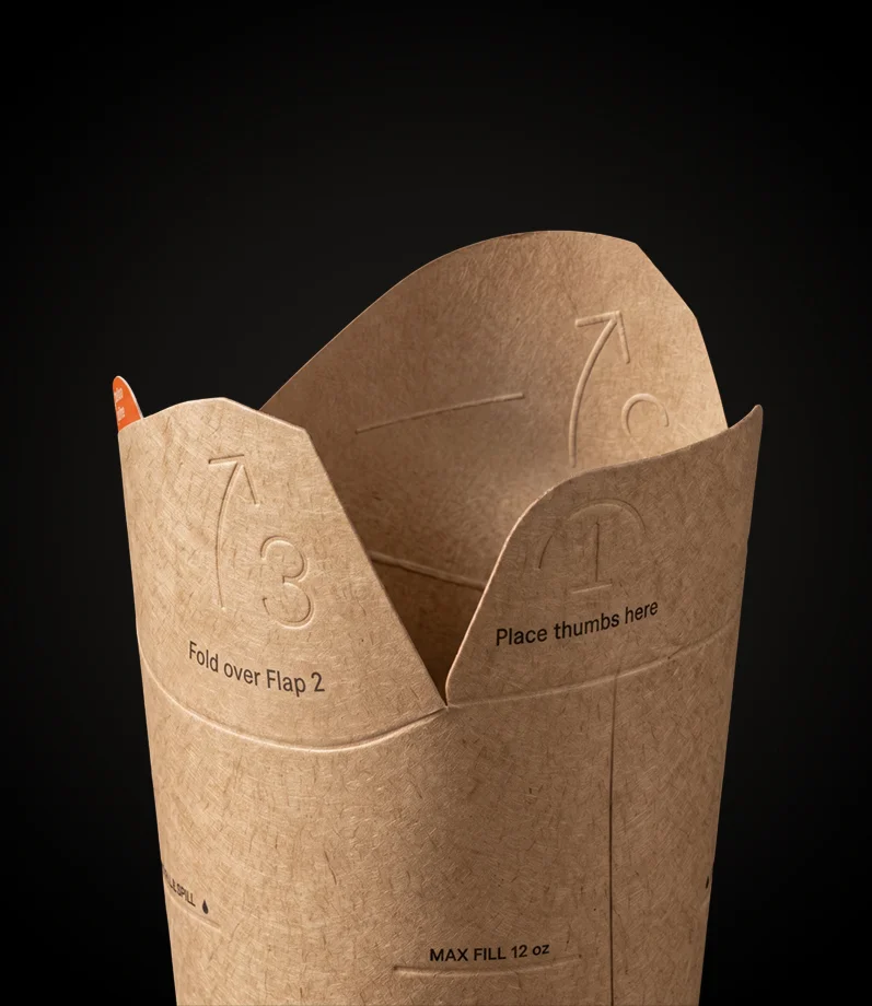 Nahaufnahme des Rands eines deckellosen Bechers aus Kraftpapier mit geprägten, nummerierten Laschen und aufgedruckten Faltanweisungen wie „Fold over Flap 2“.